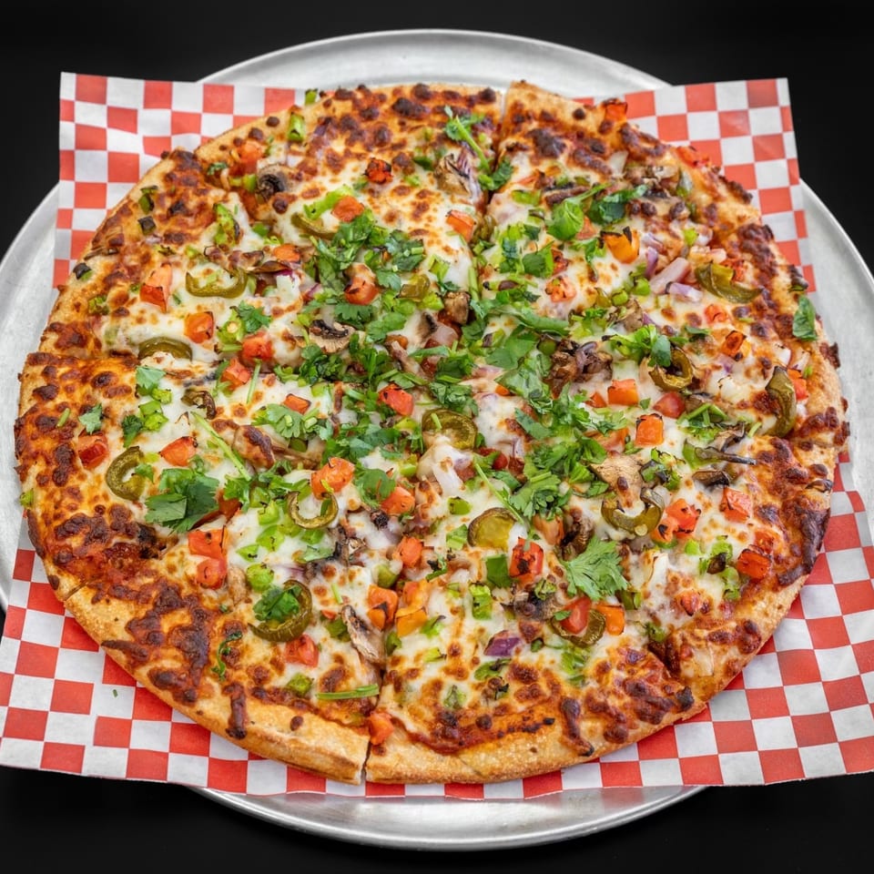 A whole pizza with melted cheese, tomatoes, jalapeños, onions, mushrooms, and cilantro on a metal tray.