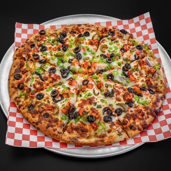 A whole pizza with chicken, black olives, green peppers, and red onions on a checkered paper.