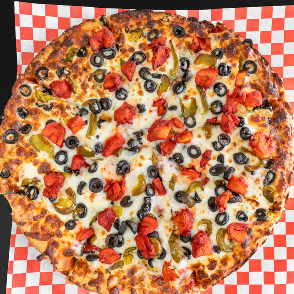 Whole pizza with red chicken, black olives, and green peppers on a checkered paper.