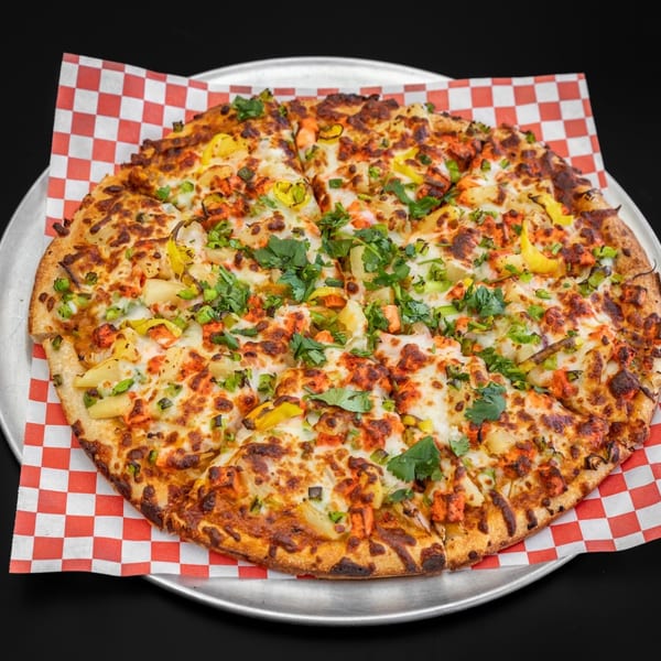 A whole tandoori chicken pizza with pineapple, green peppers, and cilantro on a metal tray.