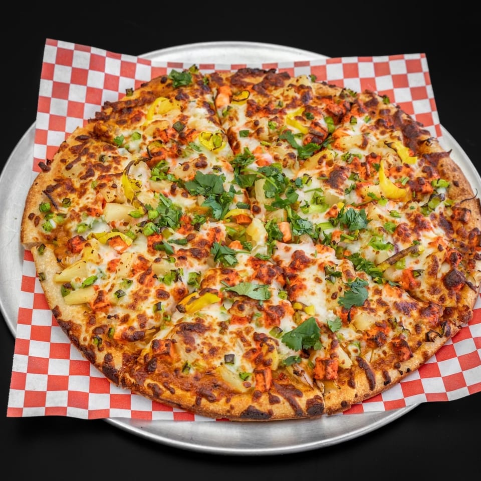 A whole tandoori chicken pizza with pineapple, green peppers, and cilantro on a metal tray.